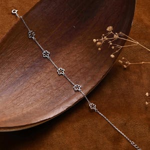 May include: A delicate silver bracelet featuring a chain with small, openwork flower charms. The bracelet is displayed on a wooden tray against a brown backdrop. The text "MICHELLELEE STUDIO Handcrafted Jewellery" is visible.