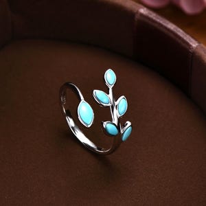May include: A silver-toned open ring featuring a branch design with five small, oval-shaped turquoise-coloured enamel leaves. The ring is displayed inside a brown circular jewellery box with the text MICHELLELEE STUDIO Handcrafted Jewellery.