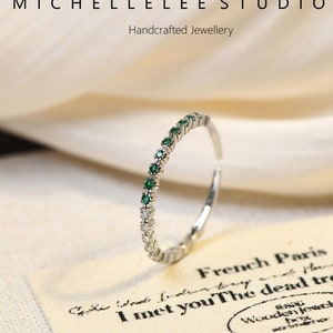 May include: A delicate silver ring with a row of small green gemstones and white diamonds. The ring is on a white fabric background with a small card that says "French Paris I met you The dead tree of spring Wooden Jewelry".