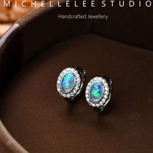 May include: A pair of oval-shaped earrings with a central blue-green opal stone, surrounded by a halo of small, clear crystals. The earrings are set in a silver-colored metal and are displayed in a brown jewelry box. The text "MICHELLELEE STUDIO Handcrafted Jewellery" is visible.