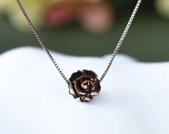 Pode incluir: Um colar de corrente de prata com um pequeno pingente em forma de rosa. A rosa é feita de metal marrom escuro com um toque de ouro.