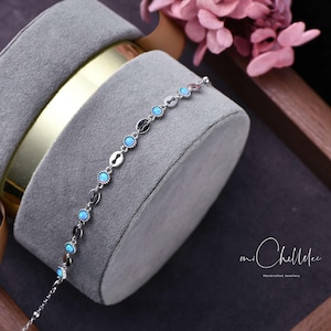 Könnte beinhalten: Ein filigranes Silberarmband mit abwechselnd ovalen und kreisförmigen Gliedern, die jeweils mit einem kleinen, runden, hellblauen Edelstein besetzt sind. Das Armband wird auf einem grauen Samt-Schmuckständer präsentiert, mit rosa Blüten im Hintergrund.