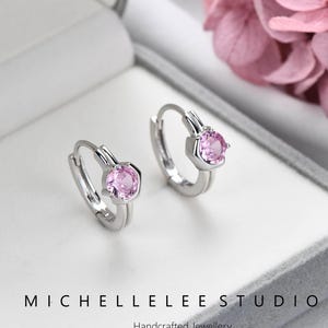 May include: A pair of silver hoop earrings with a pink gemstone center. The earrings are displayed in a gray jewelry box. The text "MICHELLELEE STUDIO Handcrafted Jewellery" is visible below the earrings.