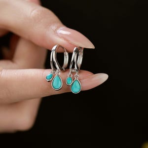 Può includere: Orecchini a cerchio in argento con pendenti a goccia turchesi. Ogni orecchino ha una piccola perla turchese sopra la goccia più grande. Il testo "MICHELLELEE STUDIO Handcrafted Jewellery" è visibile.