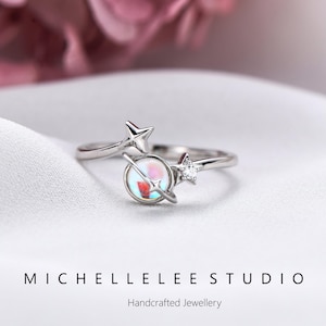 May include: A silver ring with a planet design, featuring an iridescent central stone and a small star. The ring is set against a white background. The text reads "MICHELLELEE STUDIO" and "Handcrafted Jewellery".