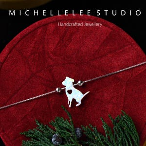 May include: A silver bracelet with a white dog charm, featuring a heart-shaped detail. The bracelet is displayed on a red surface, with the text "MICHELLELEE STUDIO Handcrafted Jewellery".