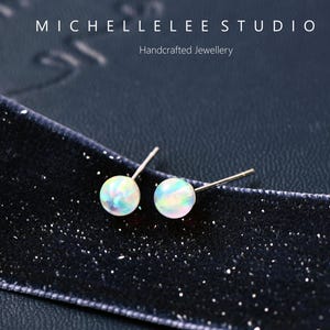 以下が含まれることがあります： 手作りのオパールスタッドピアス。各イヤリングは、銀色のポストにセットされた丸い虹色のオパールストーンが特徴です。イヤリングは、テキスト「MICHELLELEE STUDIO Handcrafted Jewellery」とともに、暗く質感のある表面に表示されます。
