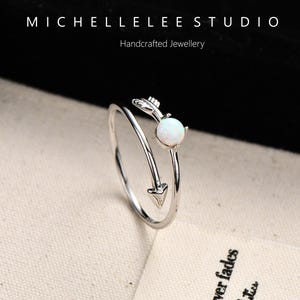 May include: A silver-colored ring with an arrow design, featuring a white opal gemstone. The ring is open and adjustable, with the arrow pointing towards the gemstone. The text "MICHELLELEE STUDIO Handcrafted Jewellery" is visible.