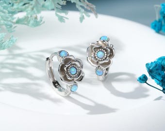 May include: A pair of silver hoop earrings featuring a floral design with small, round, iridescent blue opal stones set in the center of each petal and at the top of the hoop. The earrings have a polished metallic finish and a hinged closure.