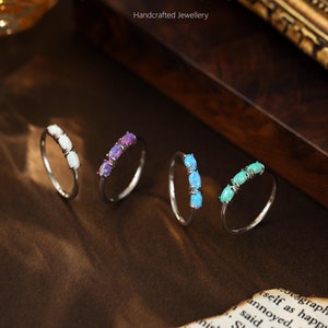 May include: Four silver rings with opal gemstones. The rings are arranged in a row on a brown surface. The opals are white, purple, blue, and green.