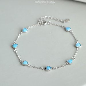 May include: A delicate silver chain bracelet with a series of small heart-shaped charms. Each charm features a light blue opal stone.