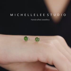 May include: A pair of small, round stud earrings with a silver setting and a green gemstone. The earrings are displayed on a person's hand. The text "MICHELLELEE STUDIO" and "Handcrafted Jewellery" are visible.