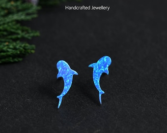 May include: A pair of stud earrings shaped like sharks, featuring a vibrant blue iridescent finish with a speckled texture. The text MICHELLELEE STUDIO Handcrafted Jewellery is visible above the earrings, which are set against a dark background.