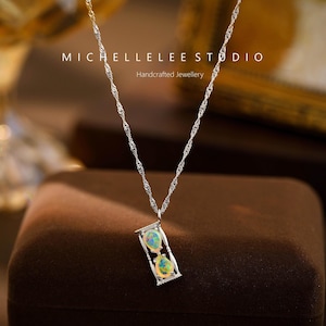 May include: A silver necklace with a pendant shaped like an hourglass. The pendant contains two iridescent, oval-shaped stones. The necklace chain has a twisted rope design. The text "MICHELLELEE STUDIO Handcrafted Jewellery" is visible.