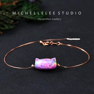 May include: A delicate rose gold bracelet featuring a cat-shaped charm with a colorful, iridescent finish. The charm displays shades of pink, purple, and blue. The bracelet is from MICHELLELEE STUDIO, Handcrafted Jewellery.