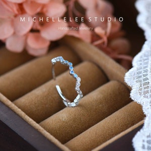 May include: A silver ring with a wavy band and blue gemstones. The ring is in a brown velvet ring box. The text "MICHELLELEE STUDIO" and "Handcrafted Jewellery" is visible on the box.