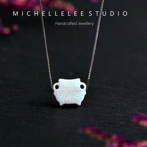May include: A delicate silver chain necklace with a small, white opal pendant in the shape of a rounded rectangle. The pendant has a slightly textured surface and two small holes on either side. The necklace is photographed against a dark background with blurred purple flowers.