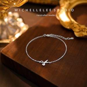 May include: A delicate silver chain bracelet with a small, clear crystal charm in the shape of a leaf. The bracelet is on a wooden surface.