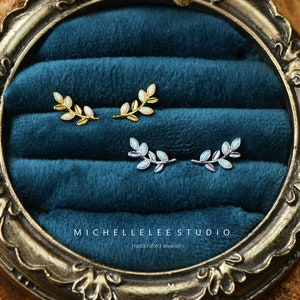 以下が含まれることがあります： 繊細な枝のデザインのイヤリングが2ペア。上のペアはホワイトオパールストーンをあしらったゴールド、下のペアはホワイトオパールストーンをあしらったシルバー。イヤリングはブルーベルベットの背景に飾られています。