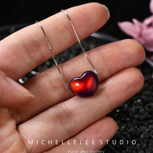 May include: A silver necklace with a heart-shaped pendant. The pendant transitions from a deep purple to a vibrant red. The necklace is held in a hand, with the text "MICHELLELEE STUDIO Handcrafted Jewellery" visible.
