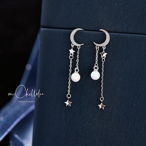 May include: Silver earrings with a crescent moon charm, a white pearl, and a star charm dangling from each earring.