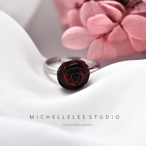 May include: A silver ring featuring a dark red rose-shaped gemstone. The ring is set against a white, silky backdrop with a blurred pink flower. The text "MICHELLE LEE STUDIO" and "Handcrafted Jewellery" are at the bottom.