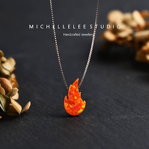 May include: A silver chain necklace featuring a flame-shaped pendant. The pendant is a bright orange with yellow and red highlights. The necklace is displayed against a dark backdrop. The text "MICHELLELEE STUDIO Handcrafted Jewellery" is visible.