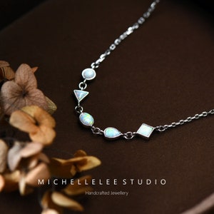 May include: A silver chain necklace with a variety of opal and white stones in different shapes, including a circle, triangle, teardrop, and square. The necklace is on a brown surface with a floral design.