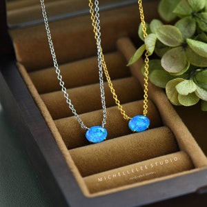 May include: Two delicate necklaces with oval blue opal pendants. One necklace has a silver chain and the other has a gold chain. The necklaces are displayed in a wooden box with the text "MICHELLELEE STUDIO Handcrafted Jewellery".