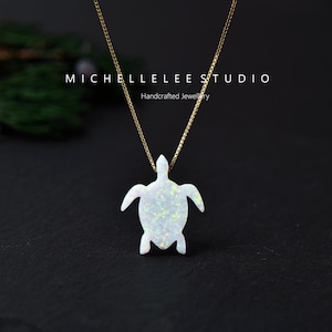 May include: A gold-colored chain necklace with a white opal turtle pendant. The pendant has a textured surface and is shaped like a turtle. The necklace is displayed against a dark background. The text "MICHELLELEE STUDIO Handcrafted Jewelry" is visible.