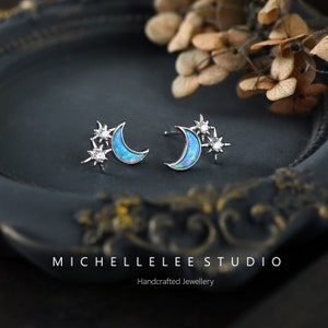 May include: A pair of silver crescent moon earrings with opal stones and clear cubic zirconia stars. The earrings are on a dark gray surface.