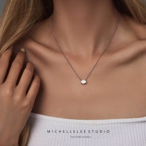 May include: A delicate silver chain necklace with a small, oval-shaped white gemstone pendant. The necklace is worn by a person with long blonde hair. The text "MICHELLE LEE STUDIO Handcrafted Jewellery" is visible at the bottom of the image.