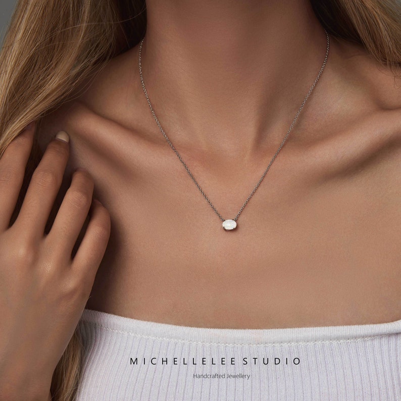 May include: A delicate silver chain necklace with a small, oval-shaped white gemstone pendant. The necklace is worn by a person with long blonde hair. The text "MICHELLE LEE STUDIO Handcrafted Jewellery" is visible at the bottom of the image.