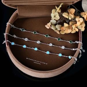Op de afbeelding: Drie zilveren kettingarmbanden met verschillende edelstenen. De bovenste armband heeft groene edelstenen, de middelste armband heeft heldere edelstenen en de onderste armband heeft turquoise edelstenen. De armbanden worden gepresenteerd in een bruine fluwelen doos met de tekst "MICHELLELEE STUDIO handcrafted jewellery".