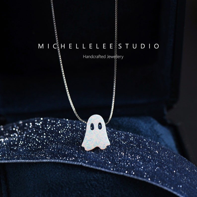 May include: A silver chain necklace with a white opal ghost pendant. The pendant has two black eyes. The necklace is in a blue velvet box with the text "MICHELLELEESTUDIO Handcrafted Jewellery".