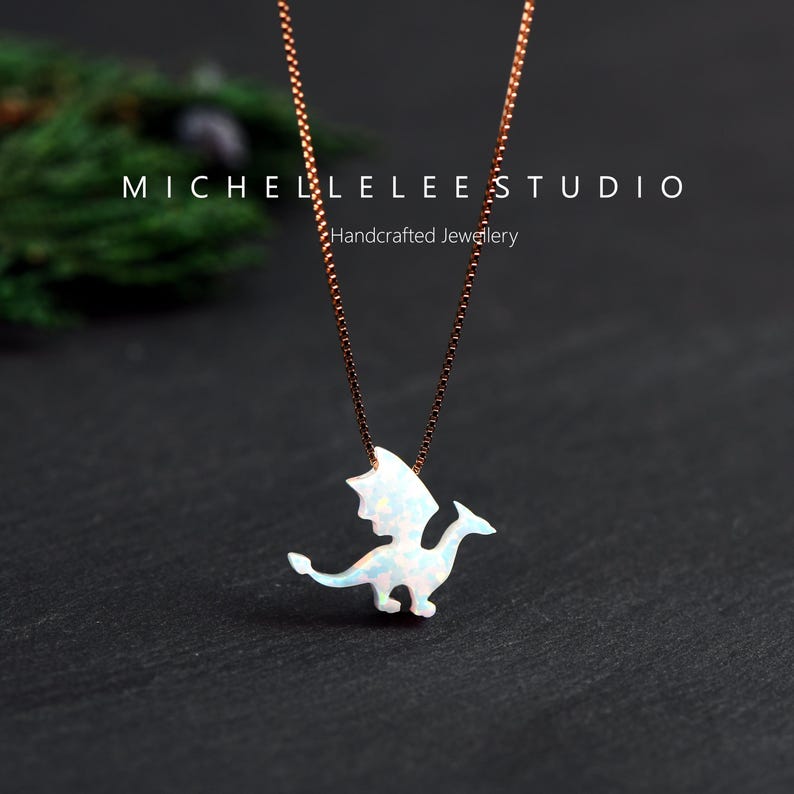 May include: A rose gold necklace with a white opal dragon pendant. The dragon has detailed wings and a long tail. The necklace is displayed on a dark surface. The text "MICHELLELEE STUDIO Handcrafted Jewellery" is visible.