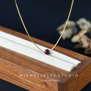 May include: A delicate gold chain necklace with a single red gemstone bead. The necklace is displayed on a wooden box with the text "Michelle Lee Studio Handcrafted Jewellery".