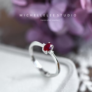 May include: A silver ring featuring a red oval gemstone and two smaller white gemstones. The ring is set against a blurred background of purple flowers. MICHELLELEE STUDIO Handcrafted Jewellery.