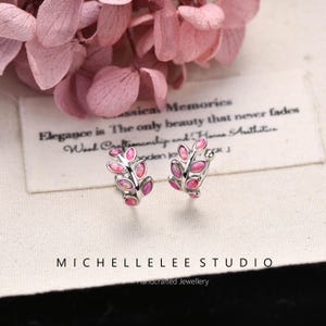 Puede incluir: Un par de pendientes tipo ear cuff plateados con forma de hoja, que presentan peque&ntilde;as piedras ovaladas de color rosa iridiscente en cada segmento. El texto MICHELLELEE STUDIO Handcrafted Jewellery aparece impreso bajo las piezas.