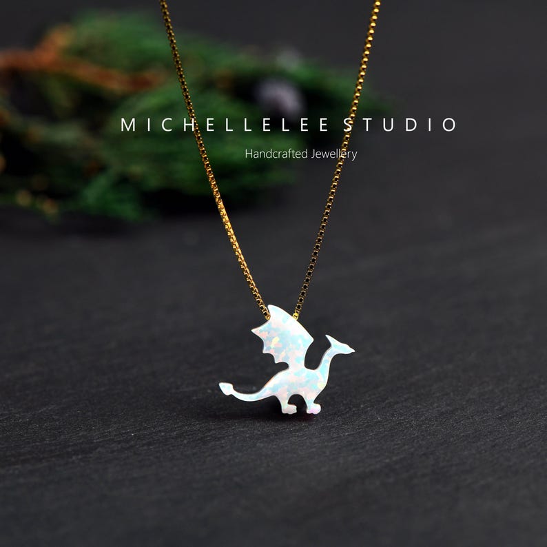 May include: A gold-colored chain necklace with a white opal dragon pendant. The dragon has outstretched wings and a long tail. The text "MICHELLELEE STUDIO Handcrafted Jewellery" is above the necklace.
