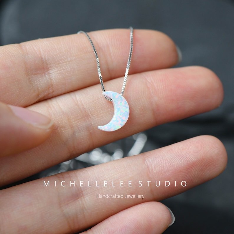 May include: A delicate silver necklace featuring a crescent moon pendant. The moon is white with a subtle, iridescent pattern. The necklace is displayed against a soft background, highlighting the handcrafted jewelry.