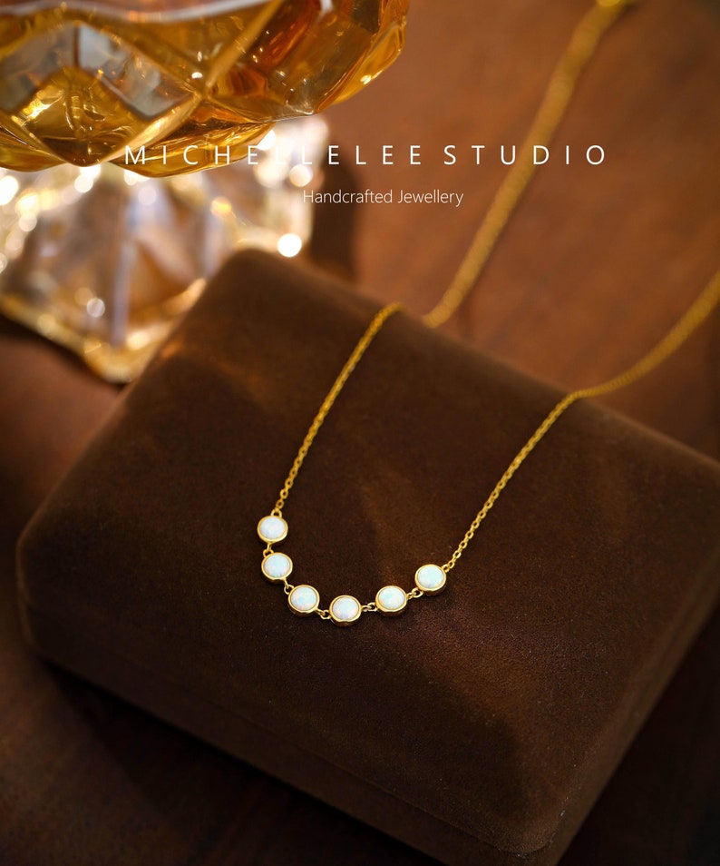 May include: A delicate gold necklace featuring a series of six small, round, white gemstones. The necklace is displayed on a brown velvet surface. The text "MICHELLE LEE STUDIO Handcrafted Jewellery" is visible in the background.