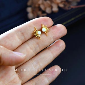 Könnte beinhalten: Goldfarbene Ohrstecker im Sonnenstrahl-Design, jeweils mit einem kleinen, runden, weißen Stein in der Mitte. Die Ohrringe werden auf einer Hand präsentiert, mit dem Text "MICHELLELEELEE STUDIO Handcrafted Jewellery" unten.