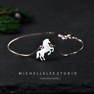 Op de afbeelding: Een zilveren armband met een witte opaal paardenhanger. Het paard staat op zijn achterbenen afgebeeld en de armband heeft een fijne ketting. De tekst "MICHELLELEE STUDIO" en "Handcrafted Jewellery" staan onderaan.