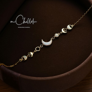 May include: A delicate gold-toned bracelet featuring moon phase charms with white mother-of-pearl inlays. The bracelet includes crescent moons, circles, and round accents. The image also shows the text "mi Chellelee Handcrafted Jewellery".