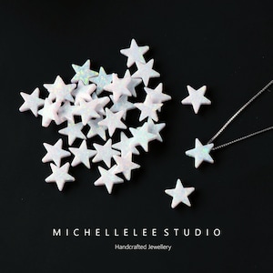 Op de afbeelding: Een stapel witte opaal stervormige kralen met een zilveren ketting en een enkele sterkraal aan de ketting. De kralen zijn verspreid over een zwart oppervlak. De tekst "MICHELLELEE STUDIO Handgemaakt sieraden" staat onderaan de afbeelding.
