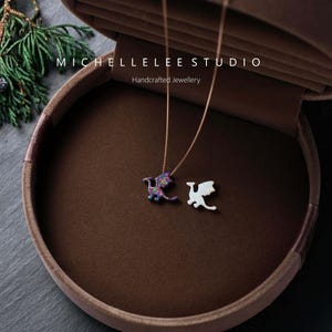 May include: Two necklaces with dragon pendants rest in a brown velvet jewelry box. One dragon pendant is white, the other is a colorful mix of purple, blue, and red. The box is open, revealing the necklaces. The text "MICHELLELEE STUDIO Handcrafted Jewellery" is visible.