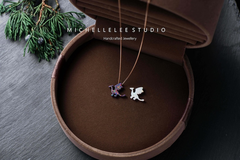 May include: Two necklaces with dragon pendants rest in a brown velvet jewelry box. One dragon pendant is white, the other is a colorful mix of purple, blue, and red. The box is open, revealing the necklaces. The text "MICHELLELEE STUDIO Handcrafted Jewellery" is visible.