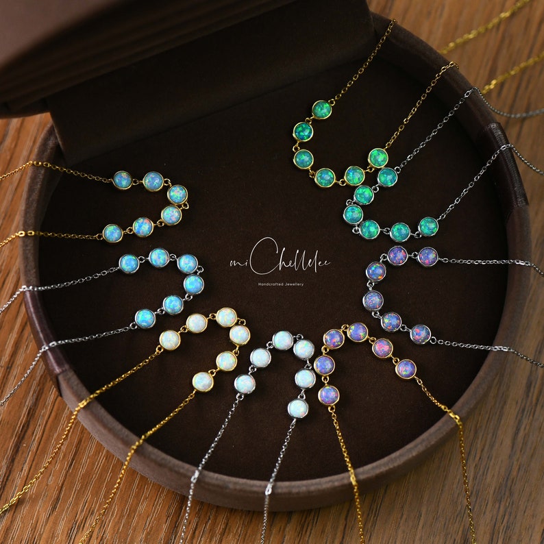 May include: A collection of delicate necklaces with opal pendants in various colors, including blue, green, and white. The necklaces are displayed in a brown jewelry box. The brand name "mi Chellelee" is visible.