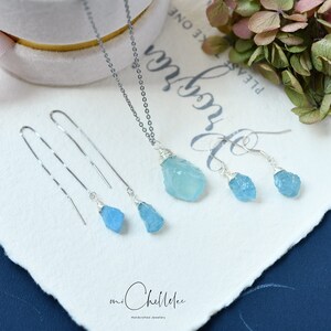 May include: A set of aquamarine gemstone jewelry featuring a necklace and earrings. The necklace has a silver chain and a pendant with a raw aquamarine stone. The earrings are silver with raw aquamarine stones.